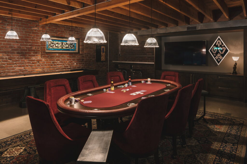 Eight-seat poker table with red velvet chairs under ribbed-glass pendants in the remodeled basement.