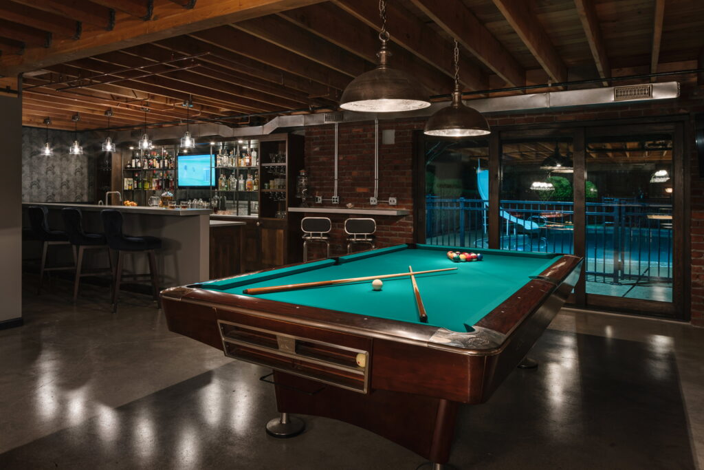 Remodeled Beaverton basement with a pool table in the foreground and a full wet bar beyond, opening onto a lit pool deck through sliding glass doors.
