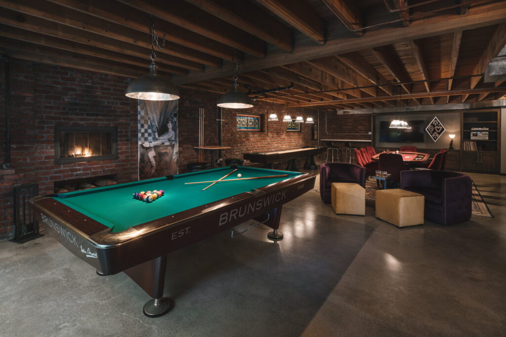 Pool table and lounge seating share the open basement floor, with a fireplace set into the brick wall at left.