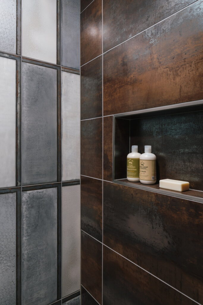 Shower detail pairing oxidized metallic-look tile with a stainless-trimmed recessed niche.