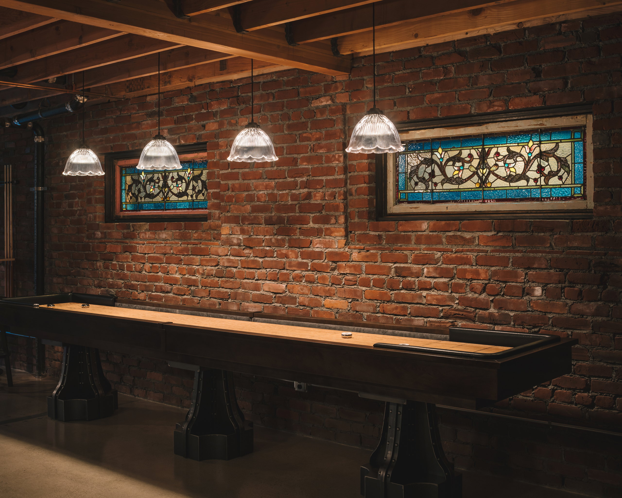 Shuffleboard table on polished concrete beneath two salvaged stained-glass windows set into an exposed brick wall.