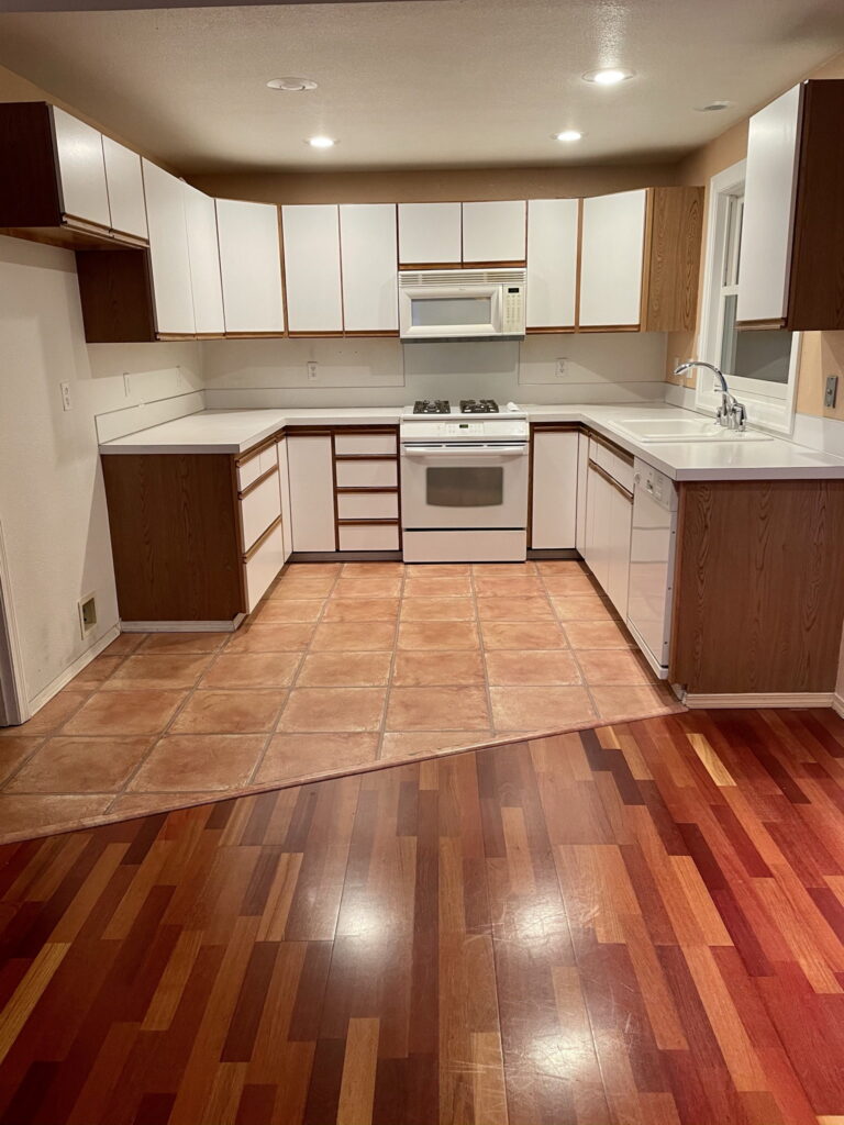 The kitchen before remodeling, with white laminate cabinets trimmed in oak, laminate counters, and terracotta floor tile.
