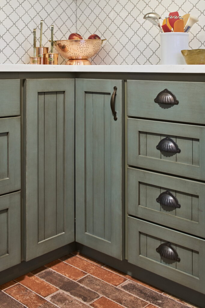 Sage-green beadboard cabinet doors and drawers with oil-rubbed bronze cup pulls and bail handles.