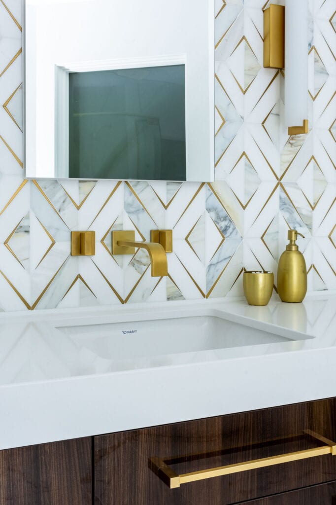 Marble tile with brass inlay in a diamond pattern, behind a brass wall-mount faucet and undermount sink.