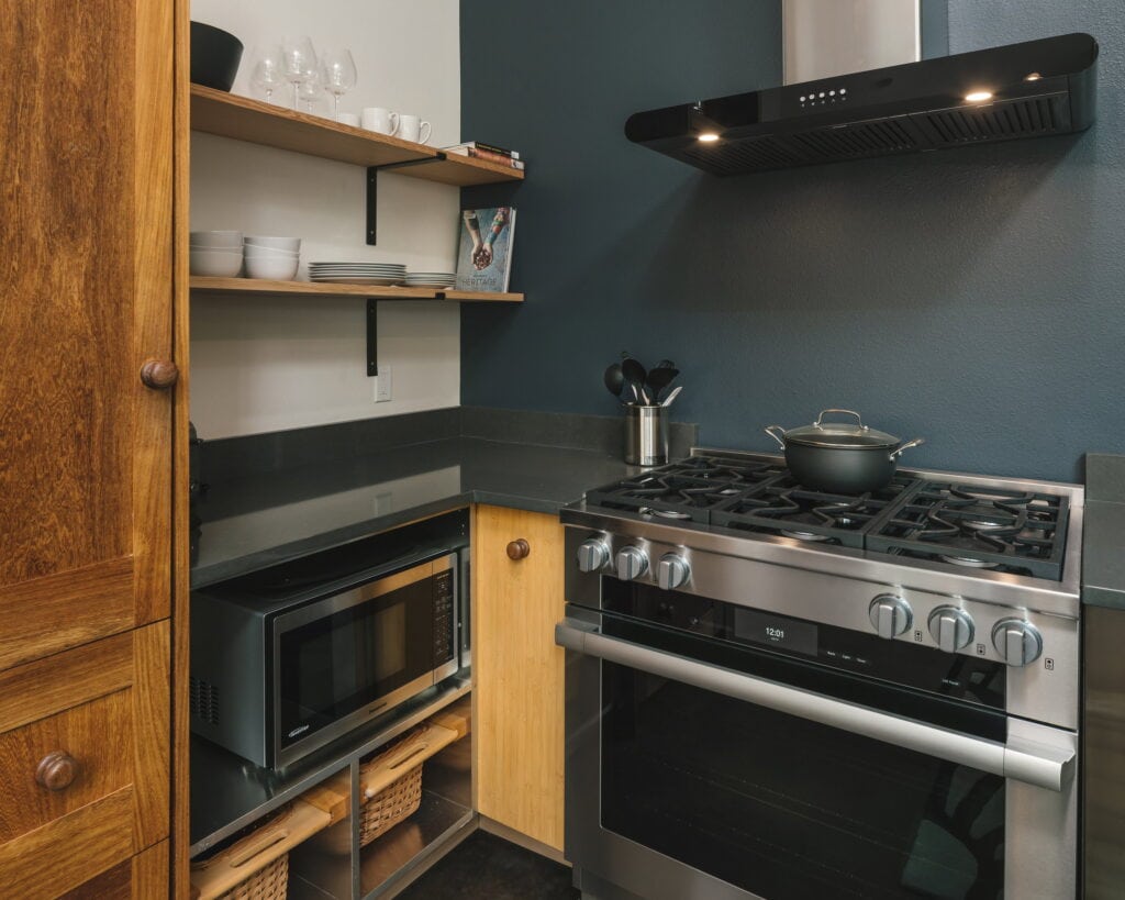 Open oak shelving and a microwave garage beside the range, against the dark blue-green wall.
