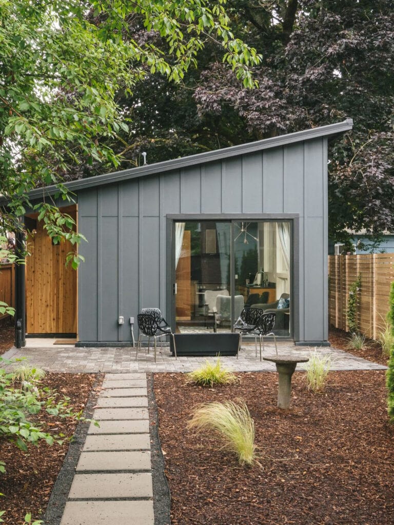 Backyard accessory dwelling unit with gray board-and-batten siding, a sloped shed roof, sliding glass doors, and a cedar-clad entry off a paver patio.