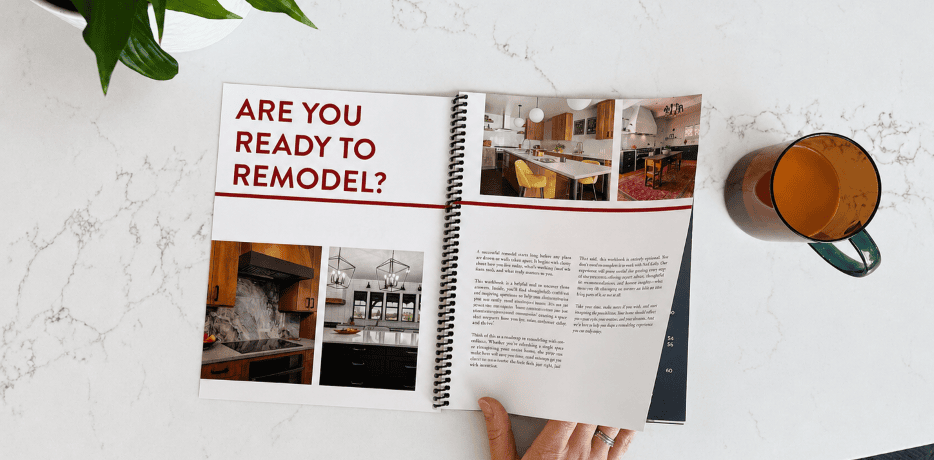 Neil Kelly’s “Are You Ready to Remodel?” workbook is open on a counter with a hand turning the page and a cup of coffee nearby
