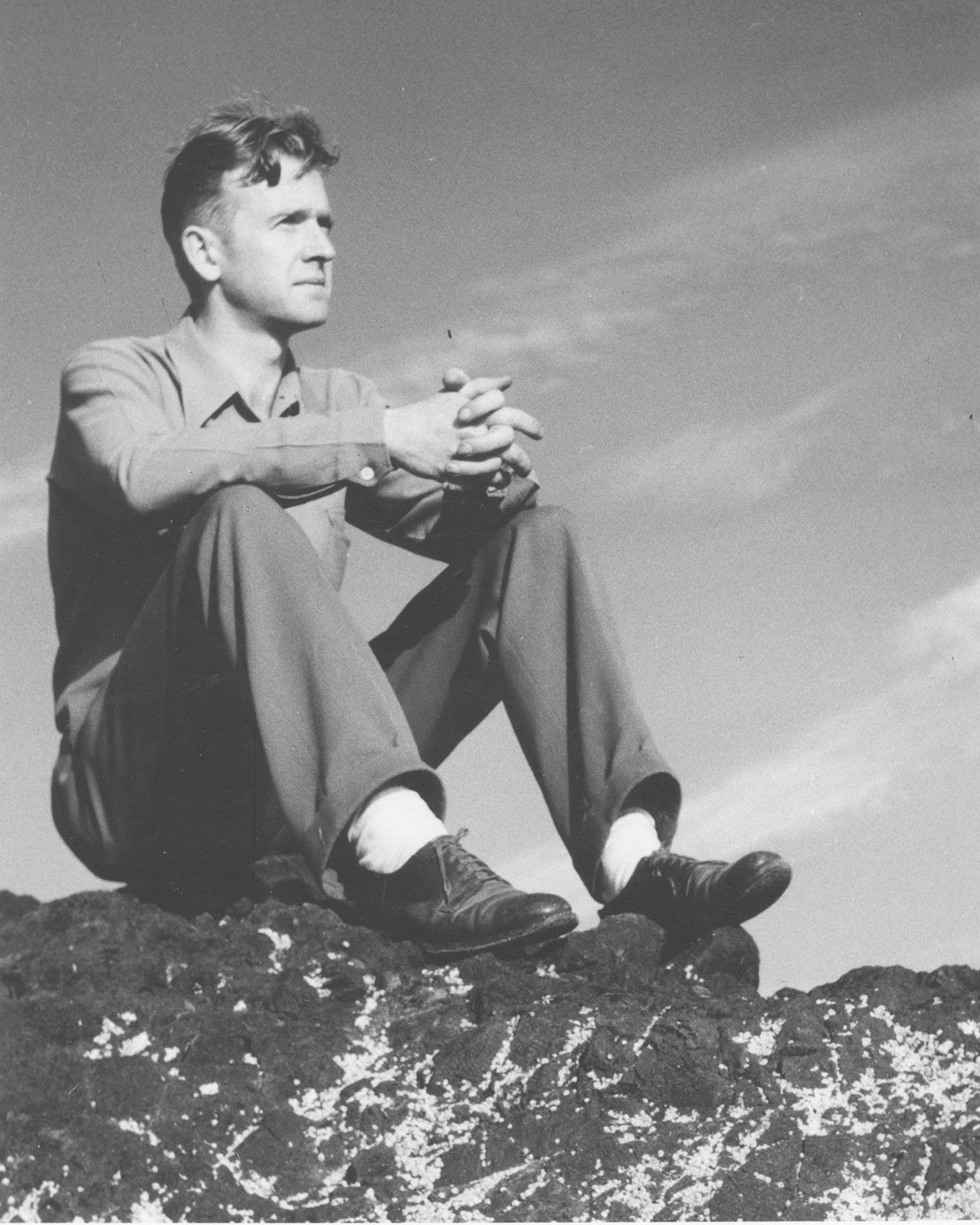 A young Neil B. Kelly sits on a rock looking into the distance with legs bent, elbows resting on his knees, and hands clasped