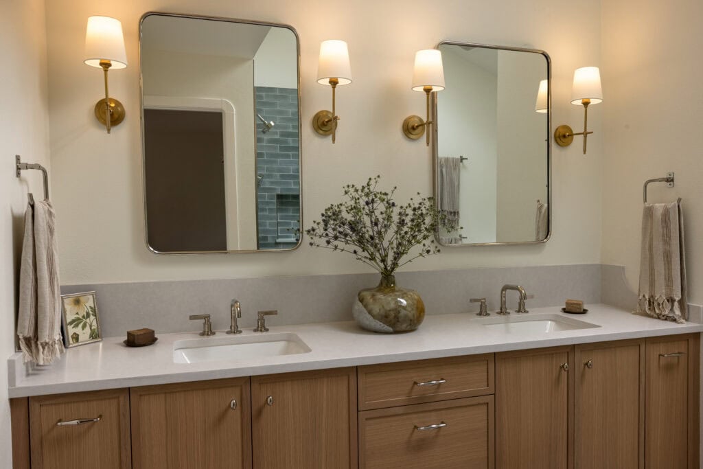 A remodeled primary bathroom with two sinks, oak cabinets, and four lamp sconces