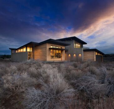 Modern custom home at dusk with large open windows with desert brush yard