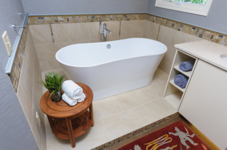 White, freestanding tub with a small, round, wooden table with a plant and three rolled towels on top.