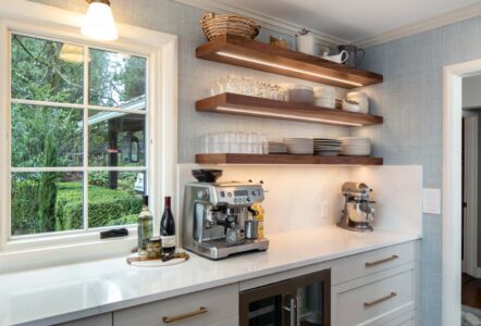 Three stacked, floating wooden shelves with kitchenware on top, mounted on the wall above a stainless steel espresso machine.