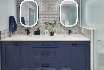 Dual LED lighted vanity mirrors above a double vanity with quartz countertops and deep blue cabinetry from Decor Cabinets.
