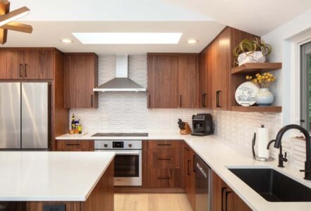 Kitchen with medium-dark wood grain cabinetry, white dimensional backsplash tile, a kitchen island with a waterfall countertop, and matte black fixtures.