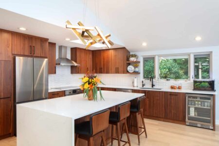 Kitchen with medium-dark wood grain cabinetry, white dimensional backsplash tile, a kitchen island with a waterfall countertop, and matte black fixtures.