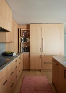A minimalist kitchen with light wooden cabinetry, sleek countertops, and a refrigerator integrated into the cabinetry, complemented by a pink rug.