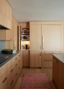 A minimalist kitchen with light wooden cabinetry, sleek countertops, and a refrigerator integrated into the cabinetry, complemented by a pink rug.
