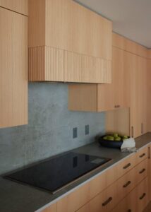 A close-up of a modern kitchen featuring light wood cabinetry, a sleek stovetop, and a stone backsplash with decorative fruit.