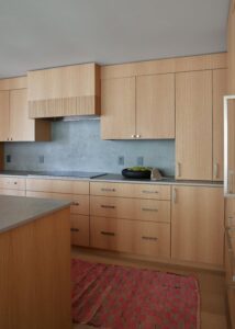 A spacious kitchen with light wooden cabinets, a smooth stone countertop, and a subtle pink rug, providing a warm and modern aesthetic.
