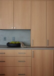 A close-up of a kitchen countertop displaying a dark bowl filled with yellow-green fruits, accented by light wooden cabinetry and a woven basket.