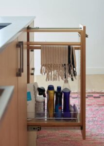 A kitchen cleaning drawer featuring hanging towels, cleaning supplies in various bottles, and a wooden rack, set against a soft pink rug.