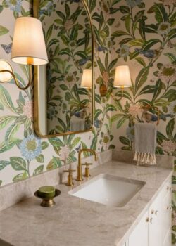 A bathroom with a white vanity and marble countertop, tropical palm wallpaper, two wall sconces, and a small mirror above the sink.