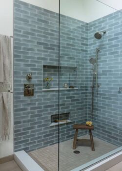 A modern glass-enclosed shower with blue brick-pattern tile walls, a built-in niche holding toiletries, a handheld shower head, and a small wooden stool inside the shower.